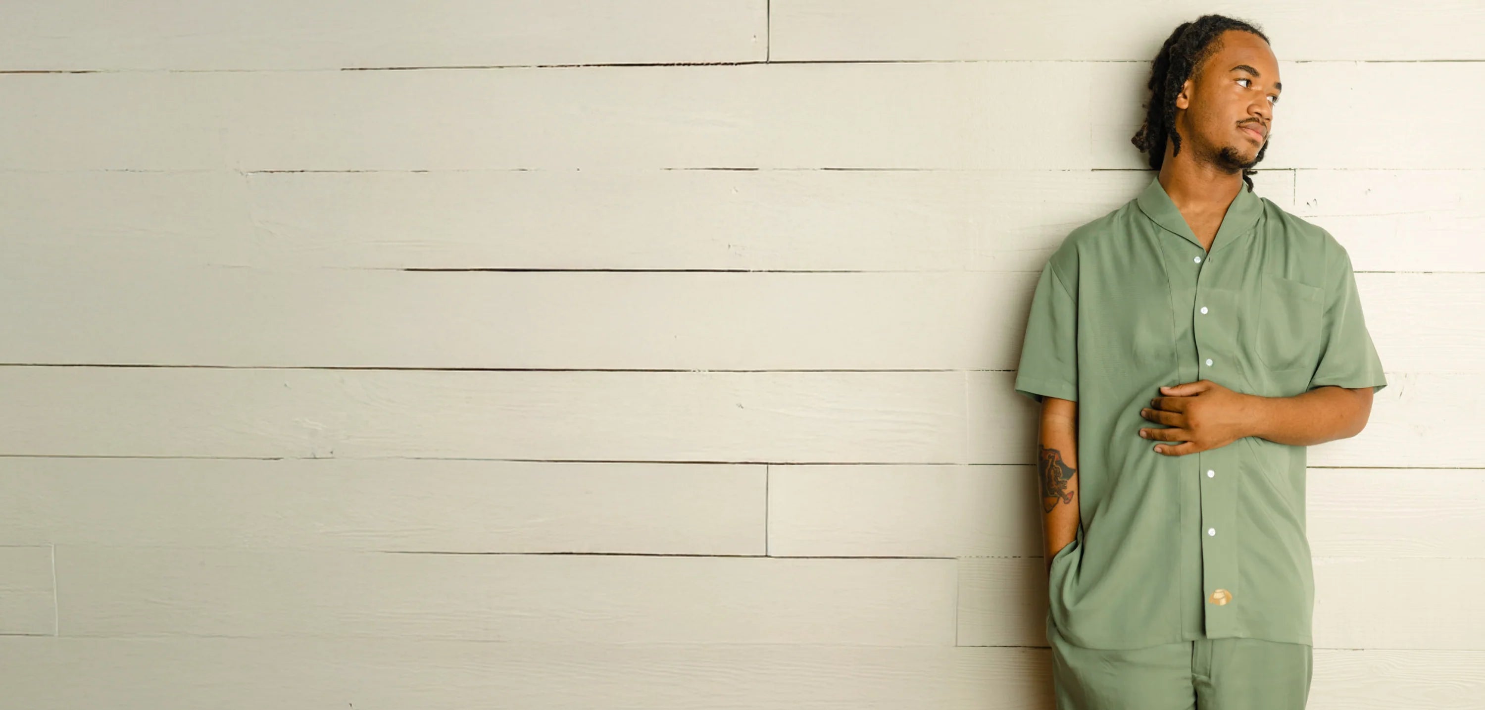 African American male with braids standing on the far right against a cream shiplap wall—head turned to the right, right arm placed in a pocket and left arm placed across his stomach—wearing a luxurious, basil green button down pajama top and pants.